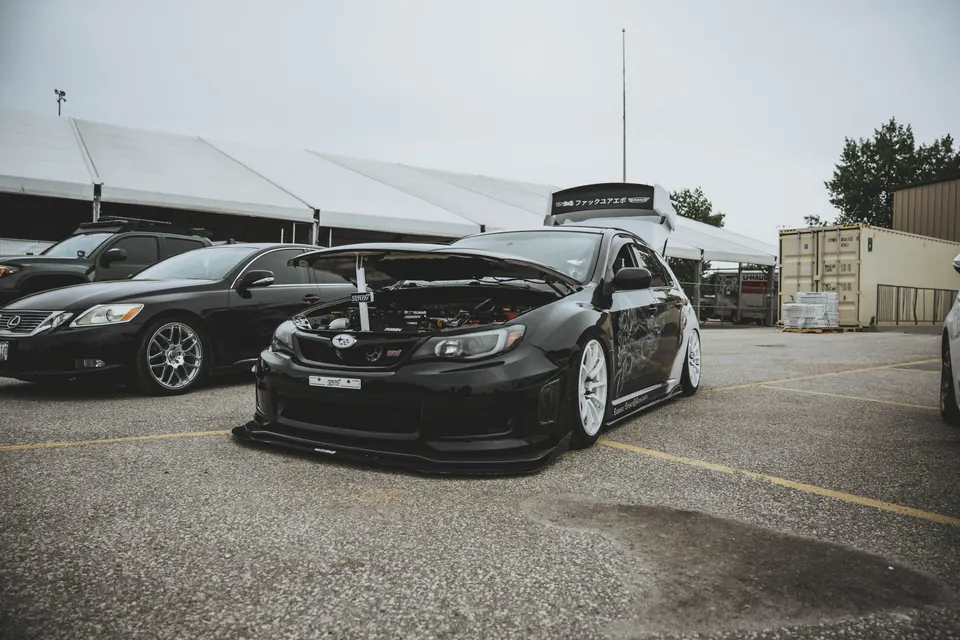 Black Subaru WRX STI hatchback with white wheels and the hood up, parked low in the show lot