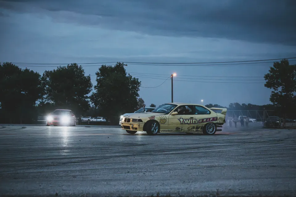 Yellow Twin Drift BMW E36 sliding with another car's headlights behind it at dusk