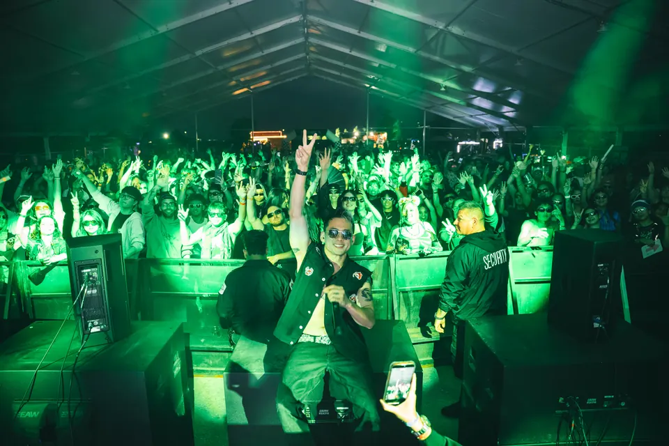 Performer throwing a peace sign on stage with a packed crowd lit green behind him